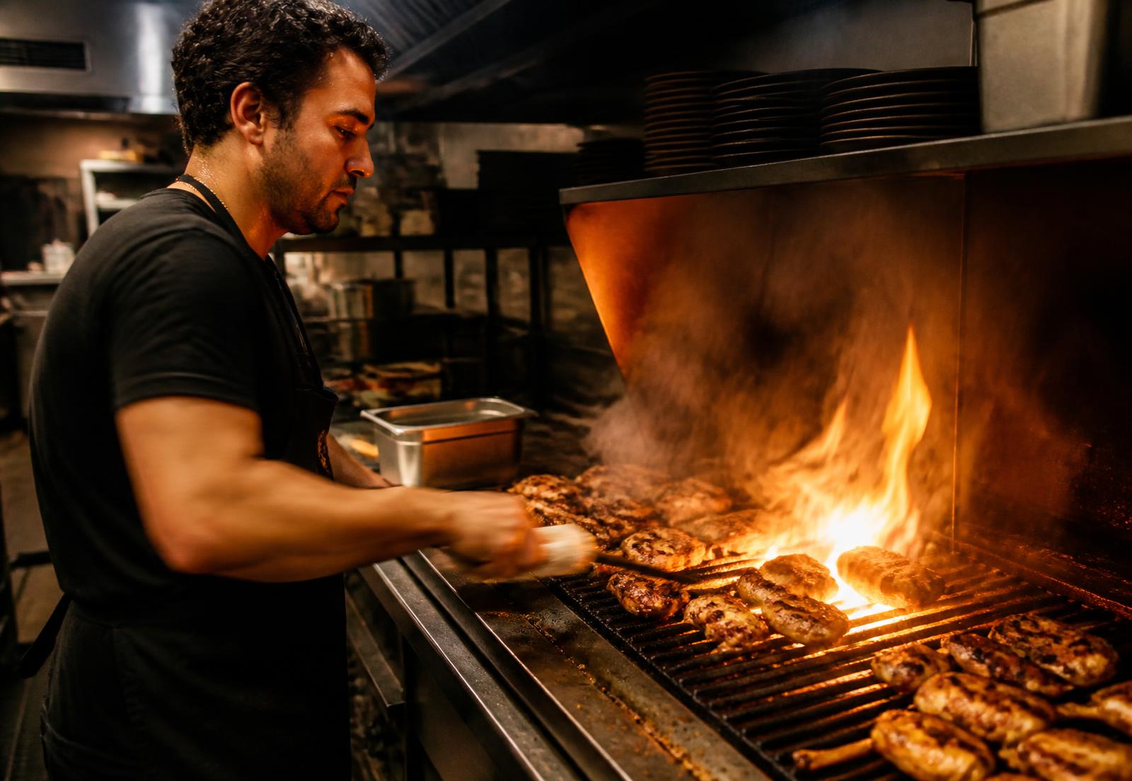 BRAVA cook working the open live-fire grill loaded with chicken, flames rising